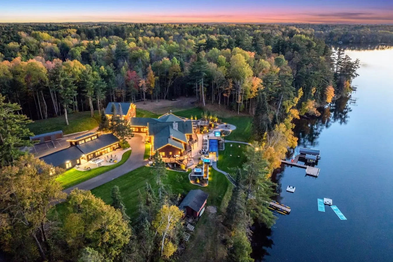 Aerial view at dusk of Long Lake Shores Estate, its lit lodge and guesthouse on a wooded point beside Long Lake